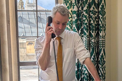 Will Forster speaking on the phone in his Westminster office, he is wearing a white shirt with the sleeves rolled up and a yellow tie.