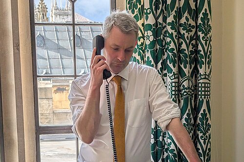 Will Forster speaking on the phone in his Westminster office, he is wearing a white shirt with the sleeves rolled up and a yellow tie.