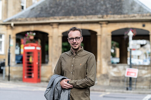 Adam Dance MP stands in the street, smiling at the camera. He is wearing a green button-up shirt and holding a grey coat. Behind him is a historic stone building with arches and a traditional red telephone box. The scene appears to be in a town centre with soft, natural lighting.