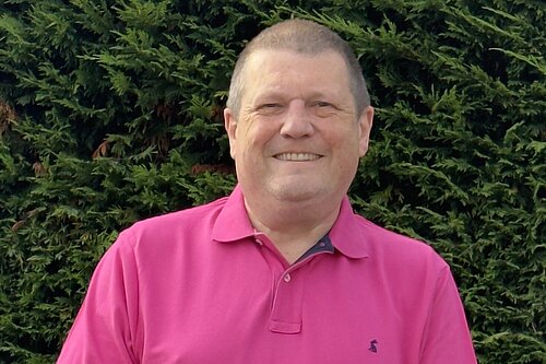 Photo of Ben Langmead smiling in a pink polo top with a hedge behind him.