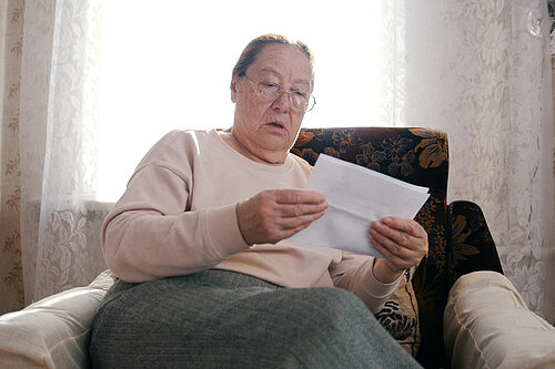 Older woman sat in arm chair looking in shock at a bill.
