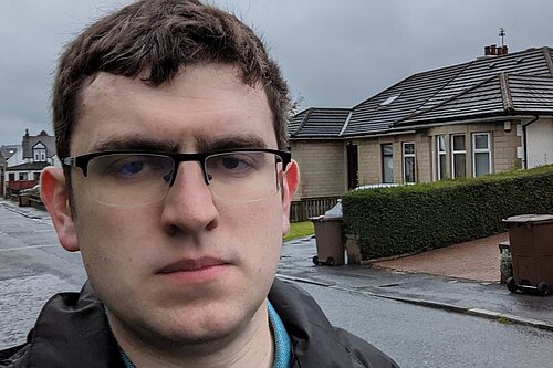 Grant Toghill, with serious looking expression, in a rain jacket with houses in the background.