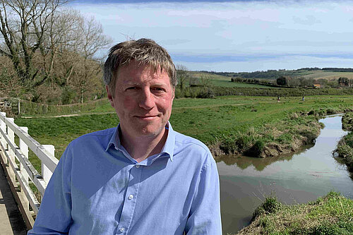 James MacCleary, in the foreground, on the South Downs Way near Alfriston, with a stream, fields, trees and blue sky behind him