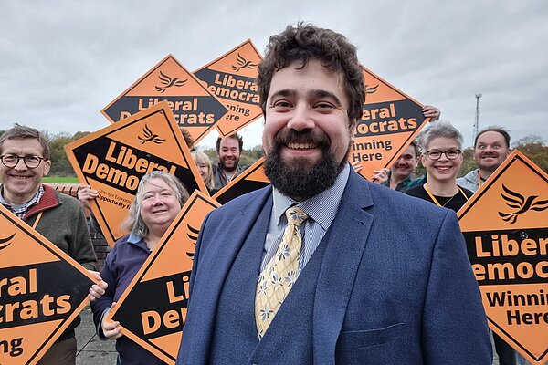 Councillor Dominic Hardwick stood infront of a group of Lib Dem campaigners holding orange Lib Dem diamonds.