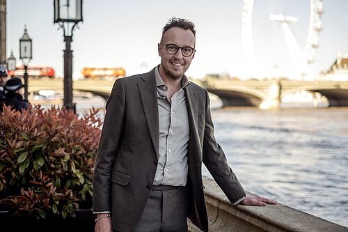 Adam Dance MP stands smiling by the River Thames in London. He is wearing a grey suit and open-collar shirt. The London Eye and Westminster Bridge are visible in the background on a bright day.