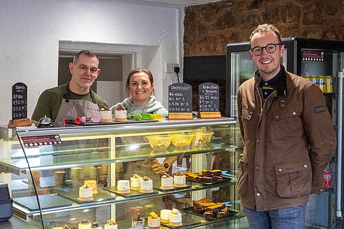 Adam Dance MP stands smiling next to two bakery staff behind a glass counter filled with cheesecakes and brownies. Chalkboard signs display the prices of the sweet treats, and the friendly setting suggests a visit to a local independent business.
