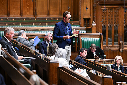 Adam Dance MP stands while others sit in a parliamentary chamber with green leather seats and wooden paneling.