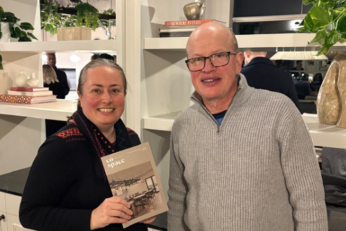 Paul and Jacqui are both smiling and standing in front of a bookcase, at the St Albans Co-Space opening event. Jacqui is holding a Co-Space leaflet.