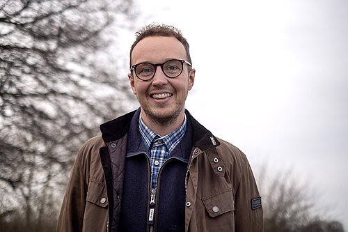 Adam Dance MP smiling outdoors on a cloudy day, wearing glasses, a checked shirt, navy jumper, and brown Barbour jacket, with bare winter trees in the background.