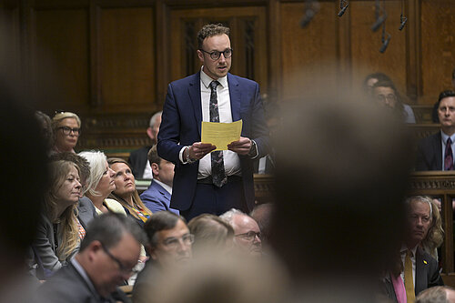Adam Dance MP stands in the House of Commons holding a yellow sheet of paper, speaking during a parliamentary session. Other MPs are seated around him, listening attentively.