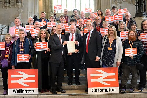 A group of people standing on the steps in Westminster Hall holding signs with the National Rail logo. There are two larger signs on the floor with the logo and with the names "Wellington" and "Cullompton" respectively. In the centre are Gideon Amos MP, Rail Minister Lord Hendy, and Richard Foord MP.