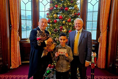 Clive Jones MP stood with local primary school student, Leo Sindhu, and the Speaker of the House of Commons, Sir Linsay Hoyle, in front of a decorated Christmas Tree. Leo Sindhu is holding his Christmas card design, which won the competition, and the Speaker is holding his large cat, named Attlee.