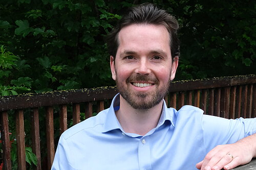 Adam Harley in a blue shirt standing with arm on top of a wooden fence of a bridge..