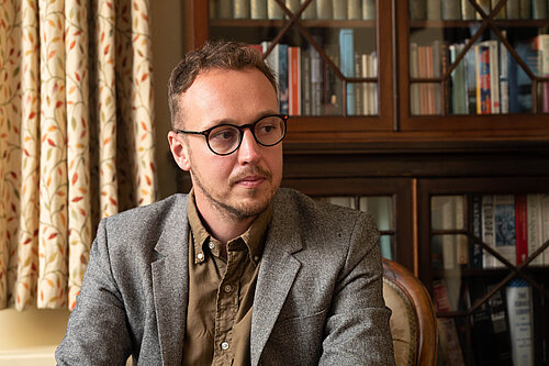 Adam Dance MP seated indoors, wearing a grey blazer and brown shirt, looking thoughtfully to the side. A bookcase and patterned curtains are in the background.