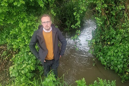 Adam Dance MP stands beside a small stream surrounded by lush green vegetation, looking up toward the camera.