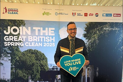 Adam Dance MP smiling while holding a heart-shaped sign that reads “Love Where You Live – Keep Britain Tidy” in front of a display for the Great British Spring Clean 2025.