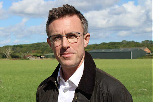 A close up of Ben Dempsey, an open field behind him and farm buildings in the background